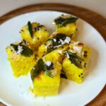 Soft, spongy yellow moong dal dhokla squares garnished with mustard seeds, fresh curry leaves, and grated coconut